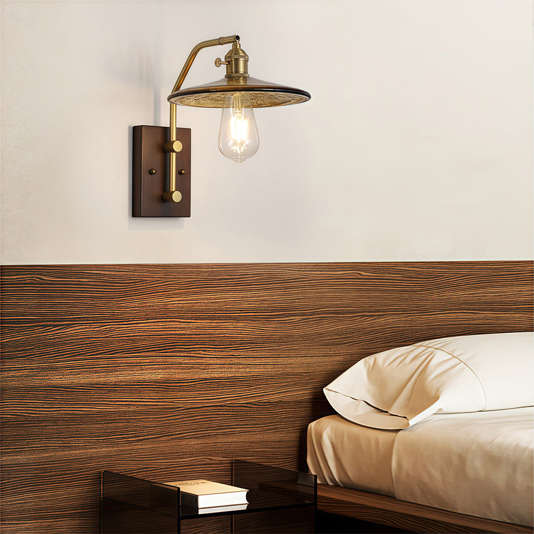 Traditional brass and glass wall lights fixture with a clear bulb, mounted on a dark wooden backplate above a bed.
